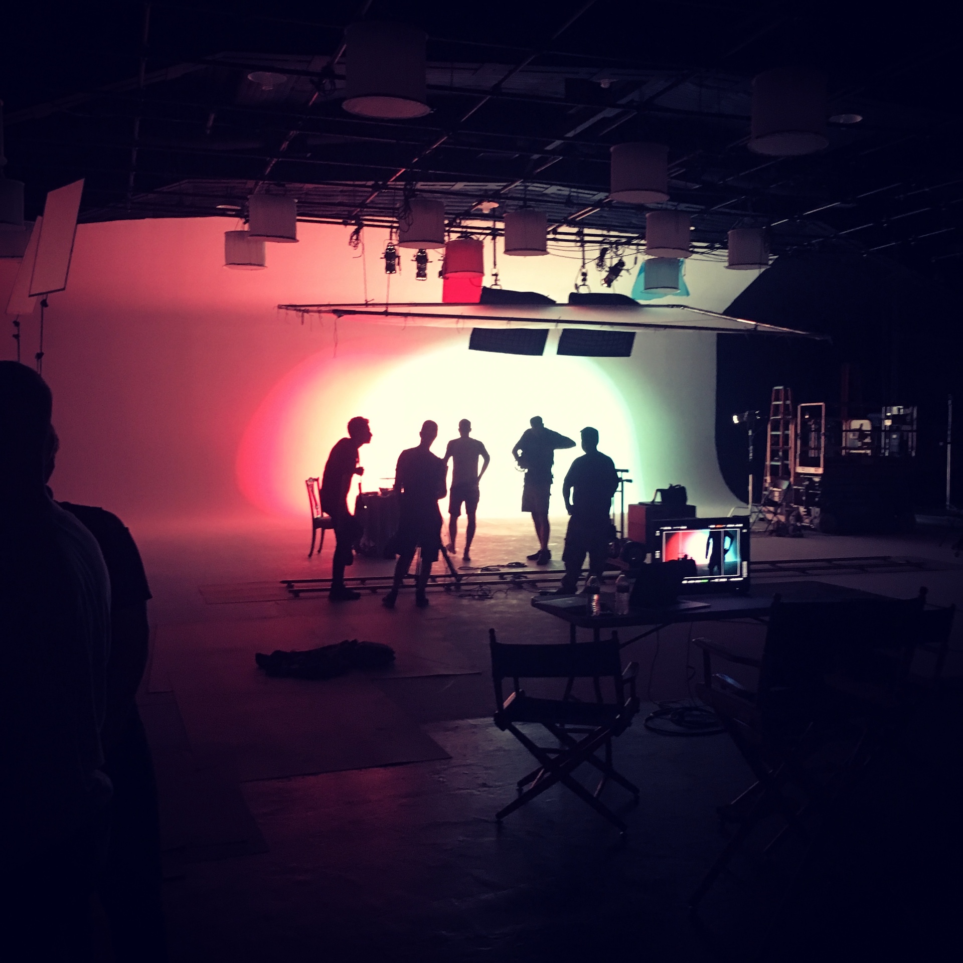 A diverse, multicultural group of young filmmakers — Black, Latina, Asian, and white women and men — gathered around a camera and monitor, lit by magenta and teal stage lights
